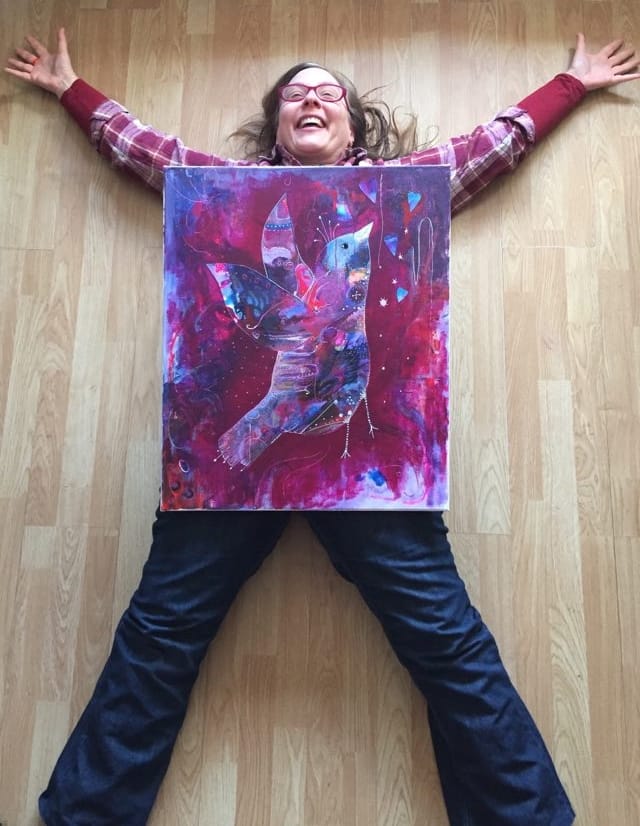 Artist lies on floor in a cross shape taking up space. Her big bold beautiful bird painting Mighty Magenta is on top of her and she is laughing with delight.